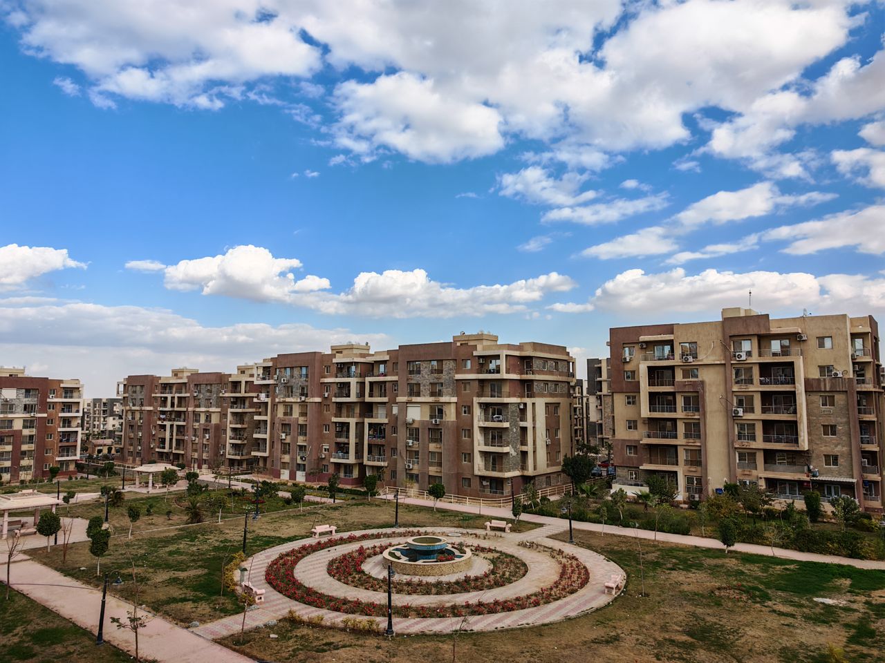 Aerial view of a modern residential complex with a central fountain under a blue sky and clouds.